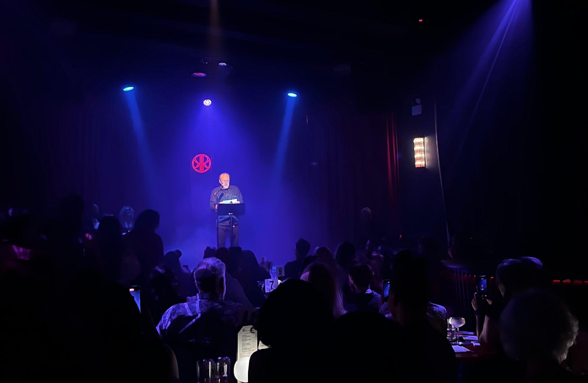 A performer reading at a lectern under blue stage lights at the Red Pavilion's Eros Unbound show